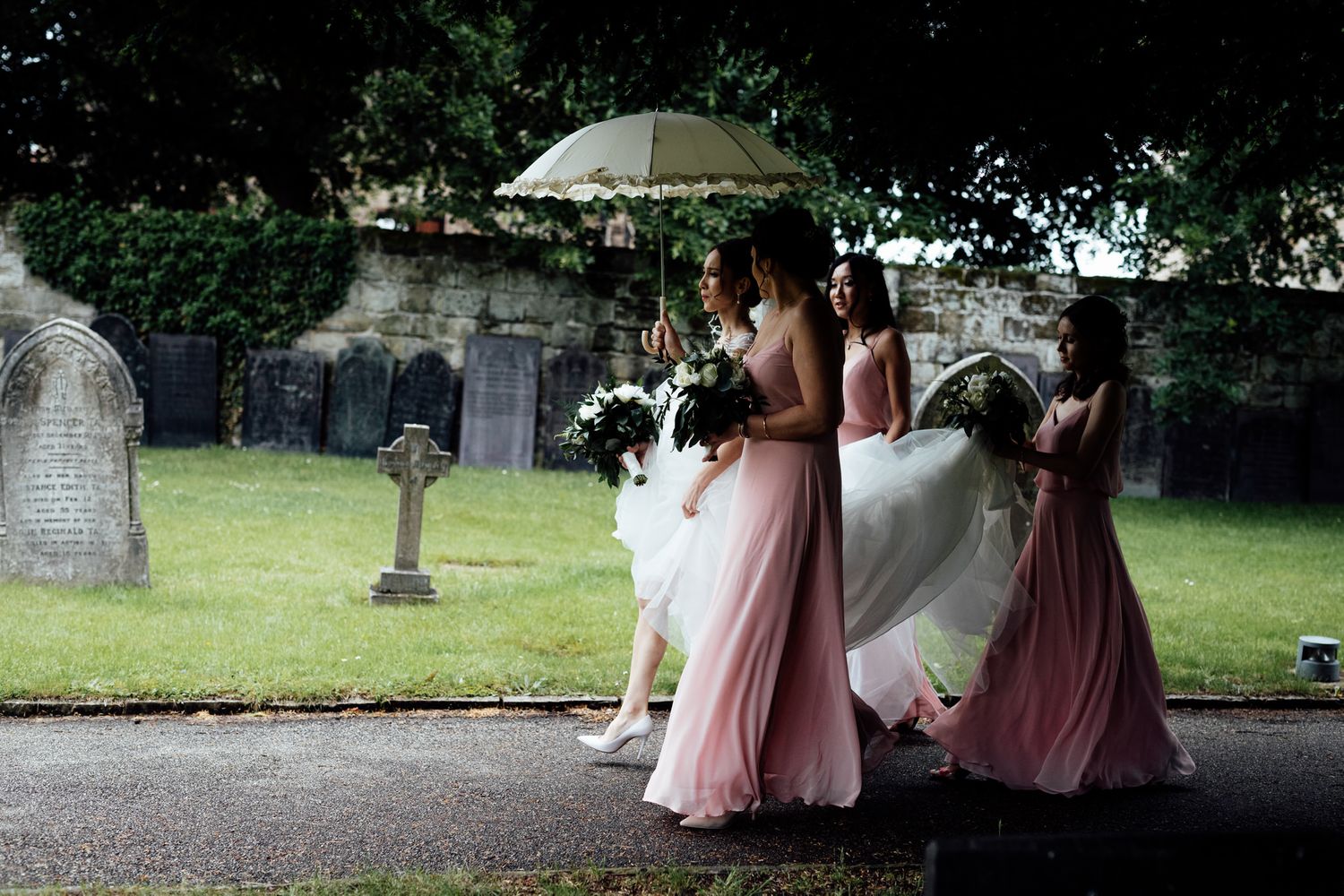 Wedding party members walking through a cemetery garden carrying pink parasols.