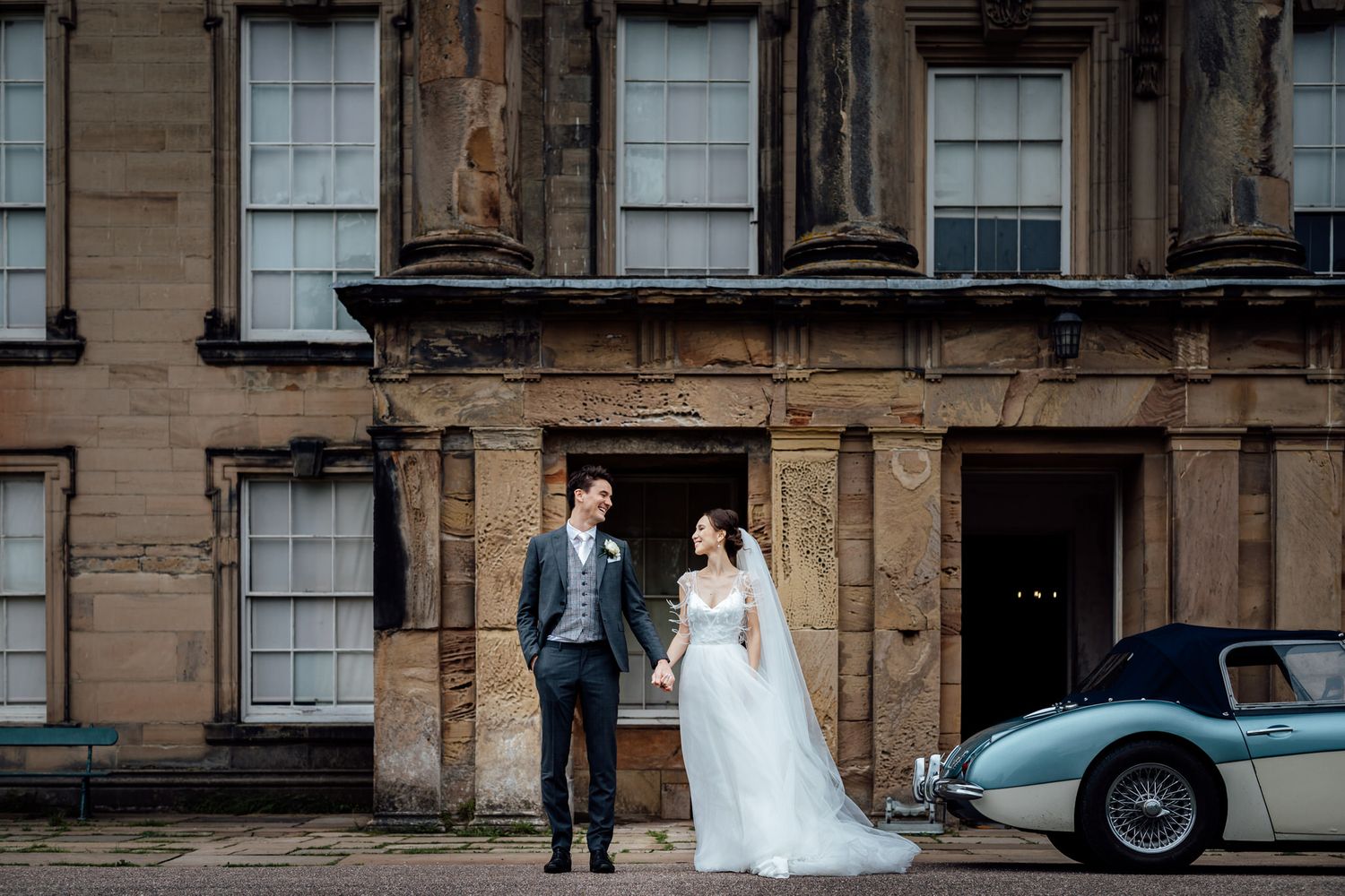 Newlyweds pose in front of historic stone building with vintage car.
