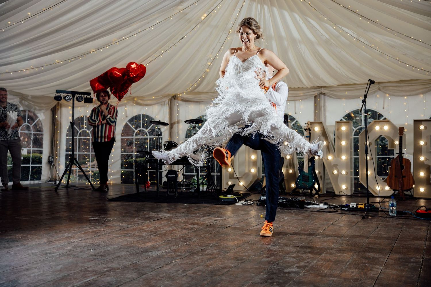 A bride in a white dress performs an energetic dance move inside a wedding reception tent.