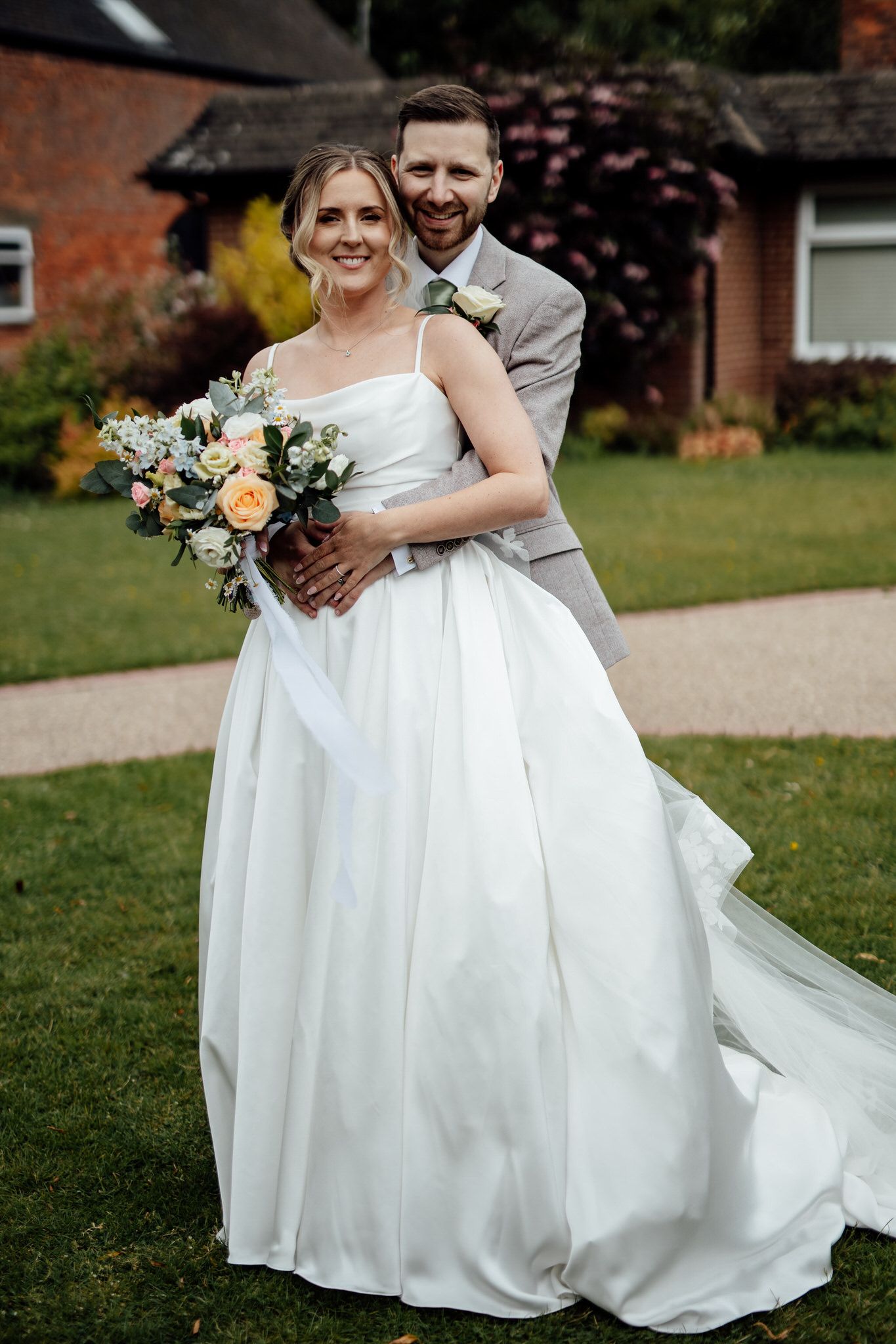 A wedding couple poses together on a lawn with beautiful floral bouquet and elegant attire.
