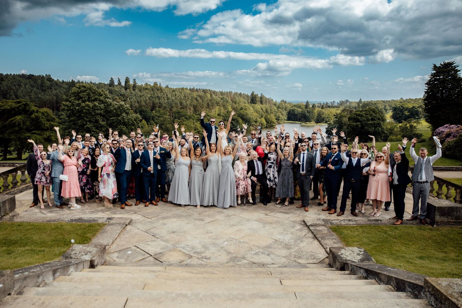 Large wedding party group photo taken outdoors with scenic mountain views in the background.