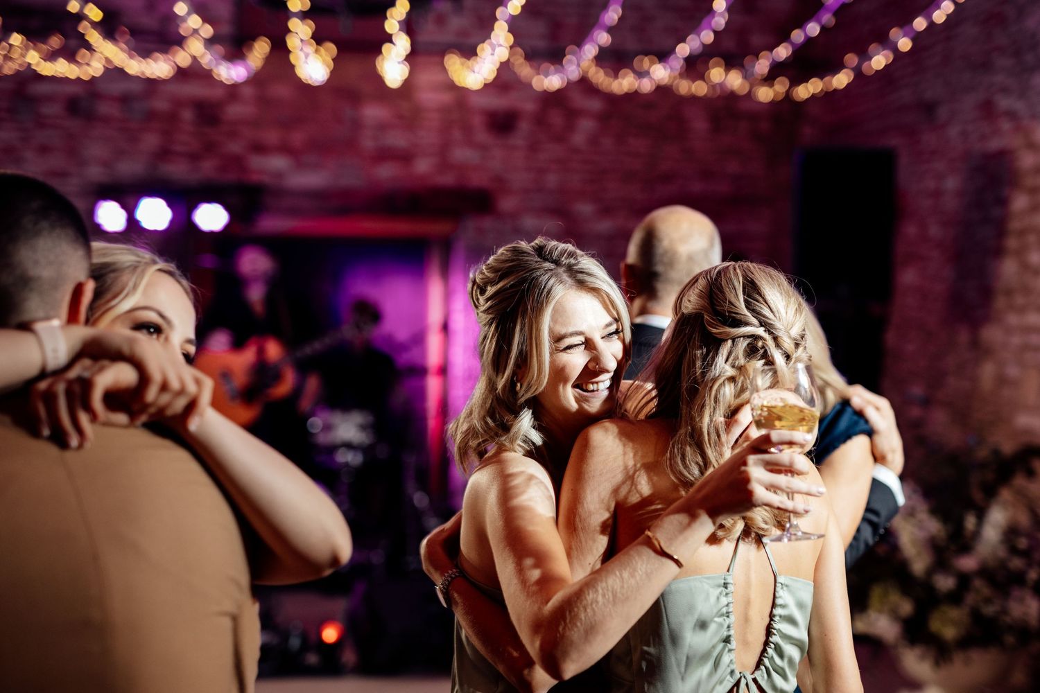 Wedding guests dancing and celebrating under warm string lights in a rustic brick venue.