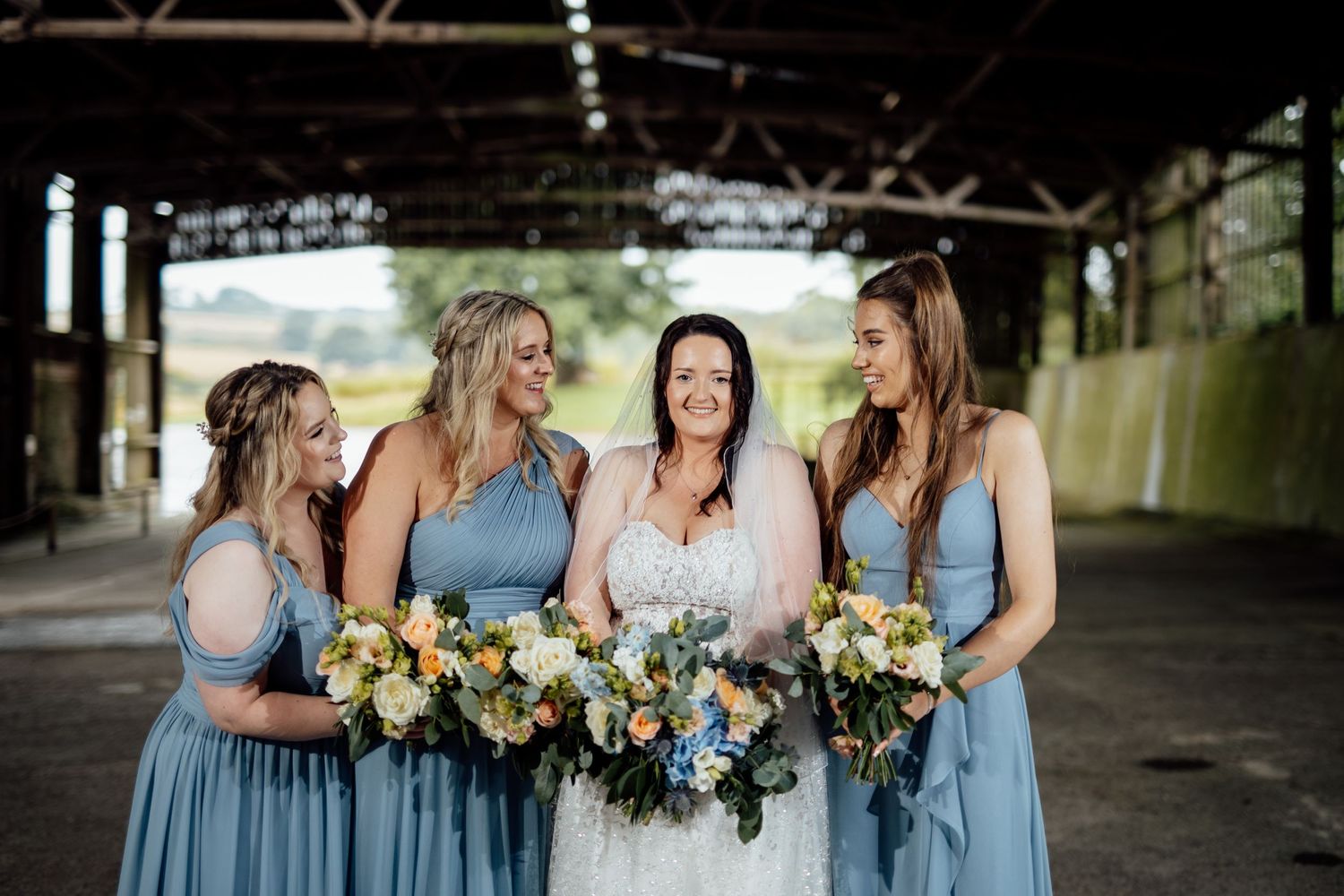 Wedding party in light blue bridesmaid dresses holding floral bouquets outdoors.