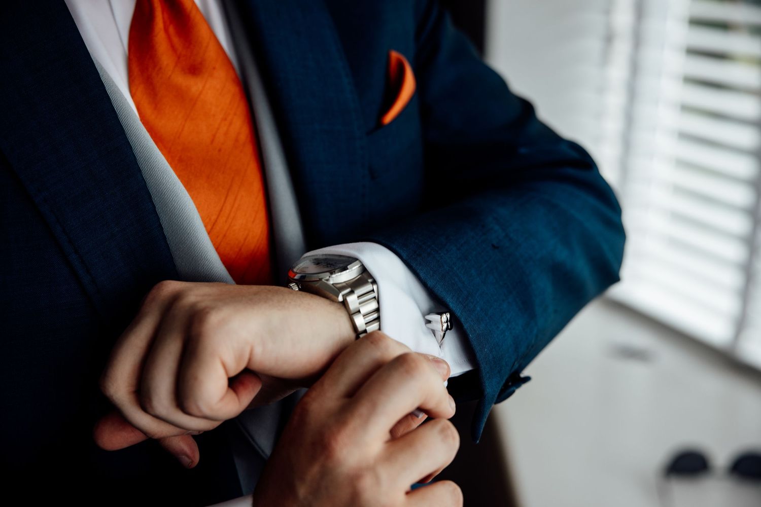 Close-up of hands adjusting a watch with orange tie and navy suit jacket visible.