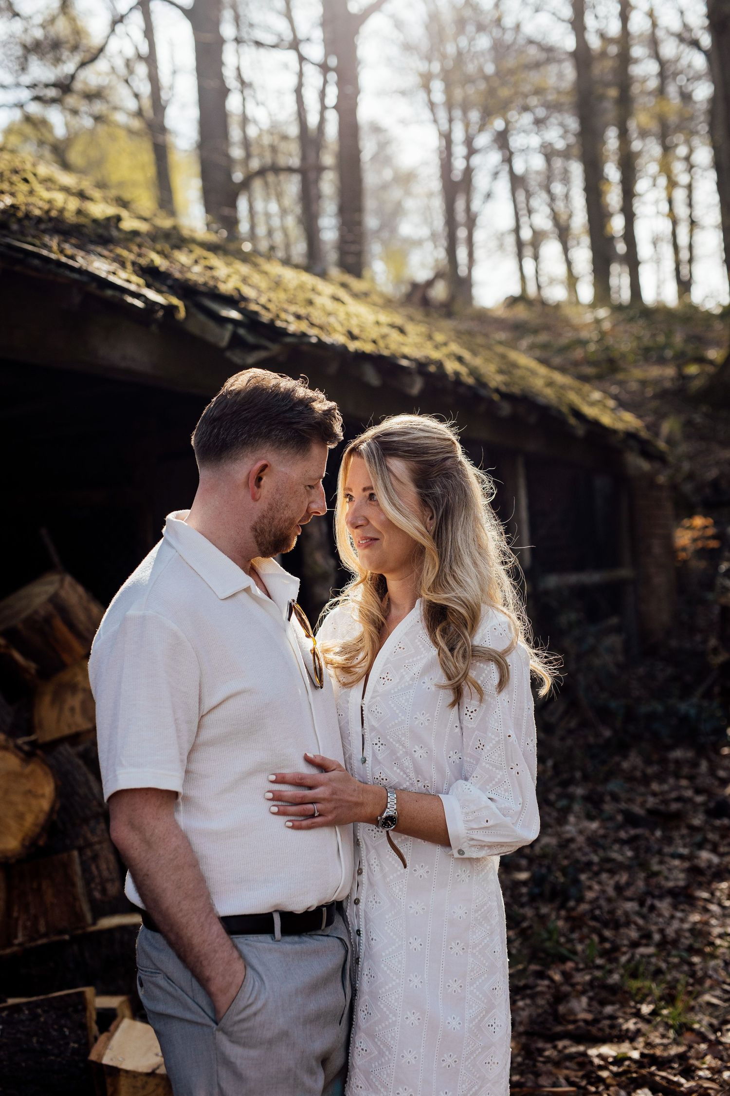 Intimate couple's photoshoot in white outfits near rustic wooden structure.