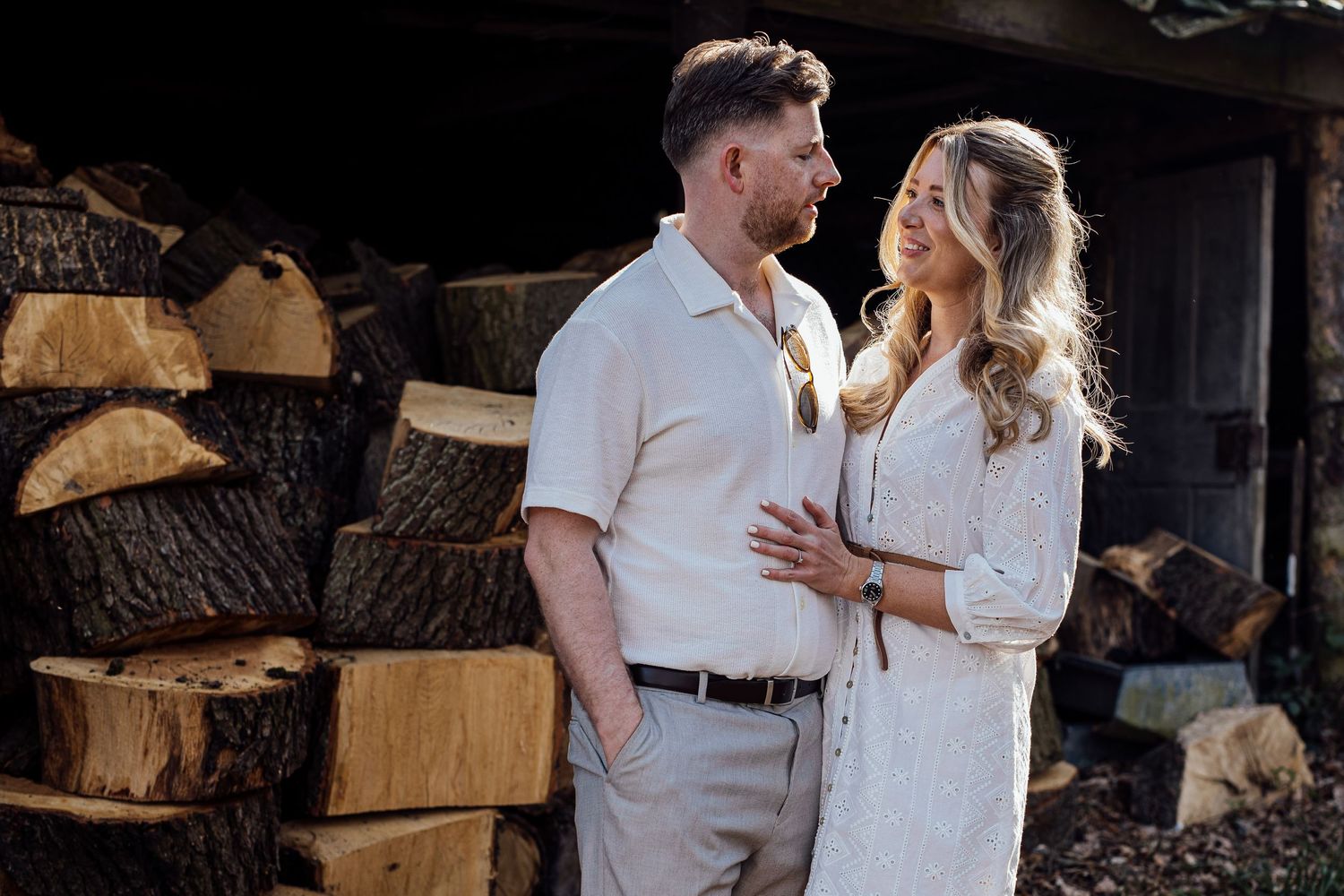 A couple in white clothing sharing an intimate moment near stacked firewood.