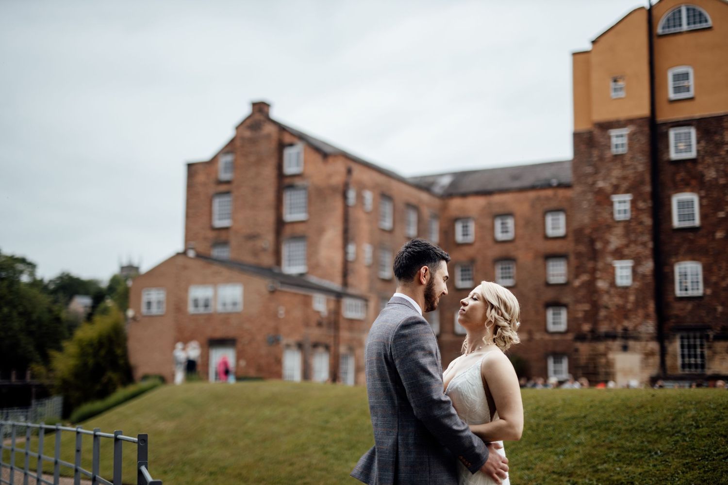 Wedding couple poses against backdrop of historic brick mill buildings on overcast day.