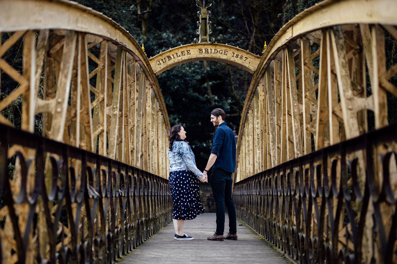 A couple walks along a rustic wooden bridge surrounded by trees and natural scenery.