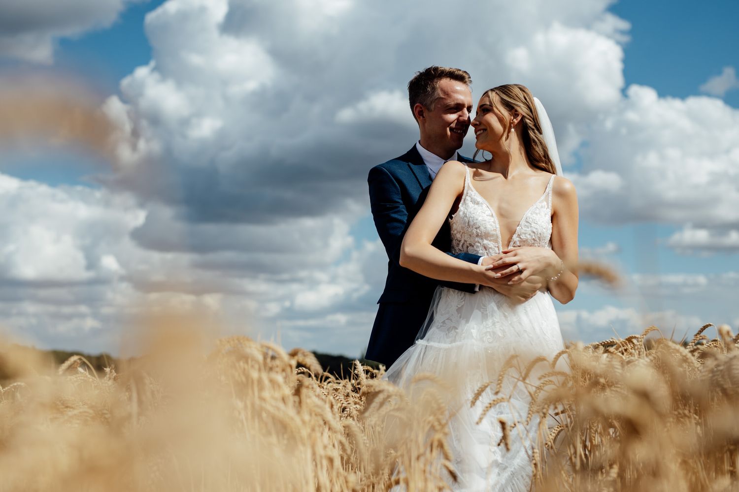 Romantic newlyweds embrace in a sun-lit wheat field under blue sky with white clouds.
