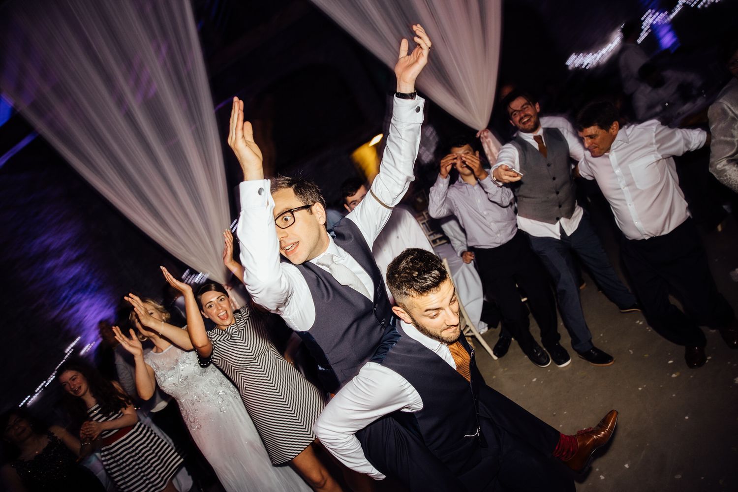 Silhouettes of wedding guests dancing against white drapes and colored lighting.