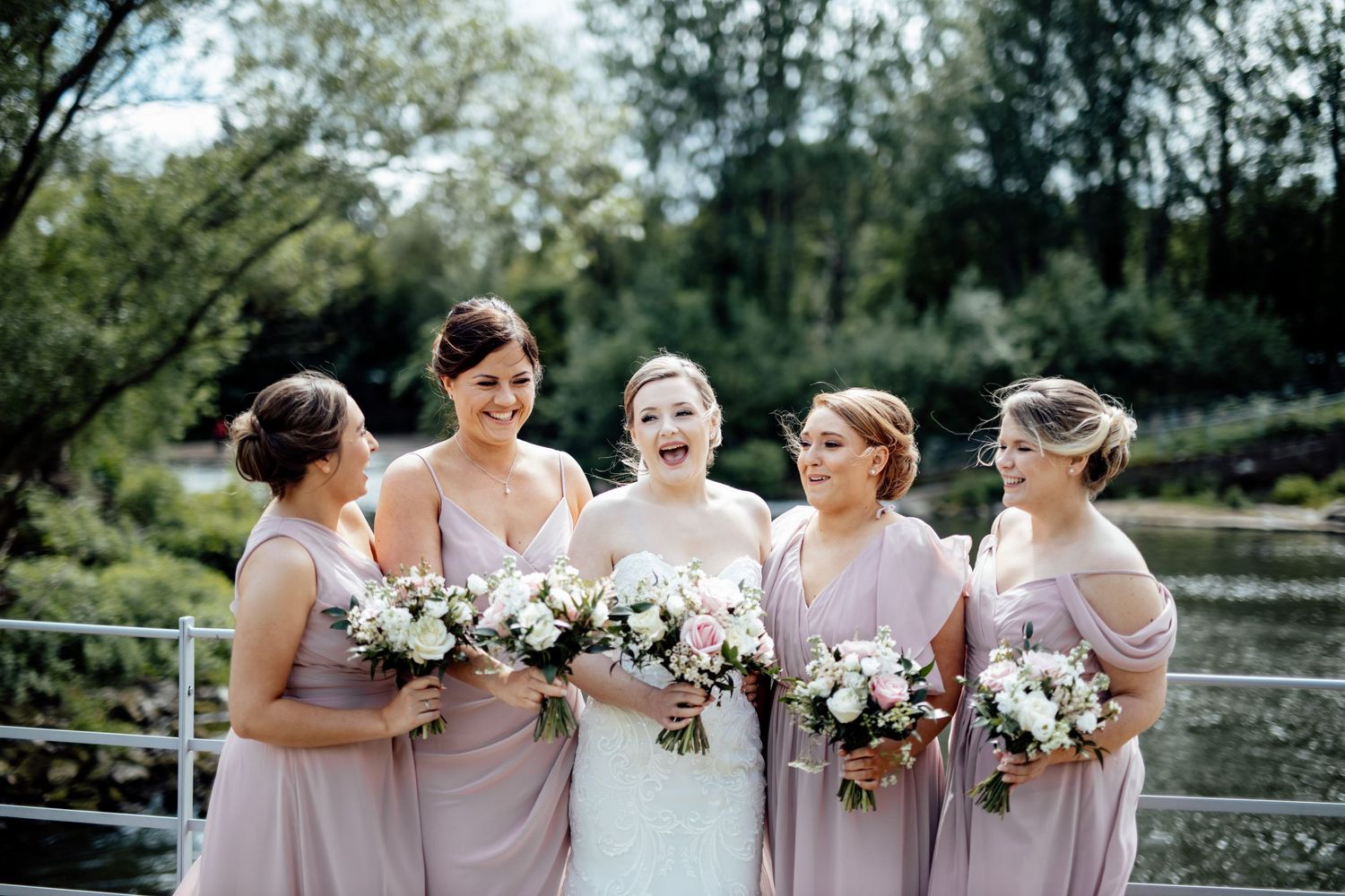 Bridesmaids in blush pink dresses pose together outdoors with floral bouquets.