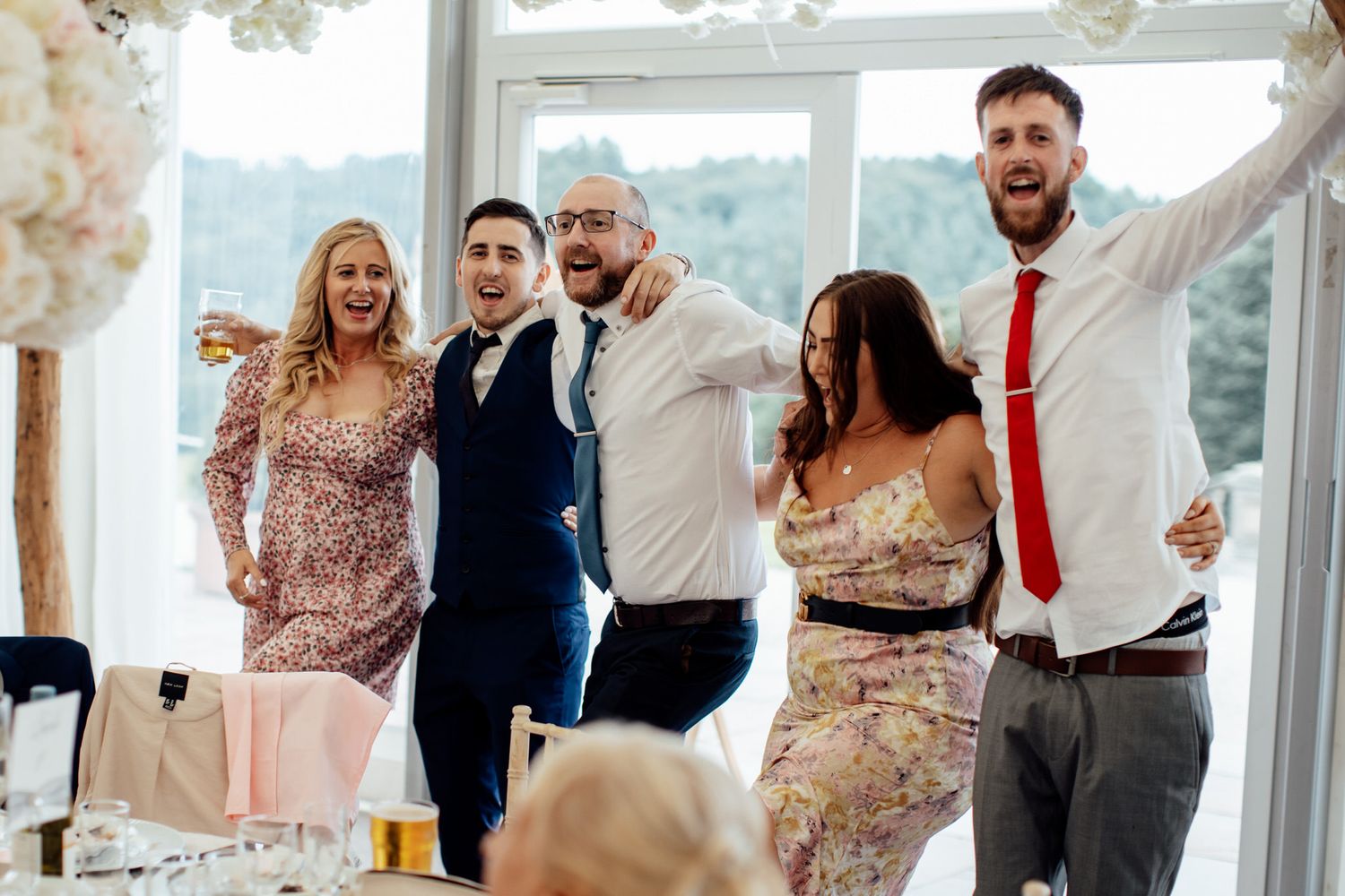 Wedding reception celebration in bright sunlit room with large windows overlooking scenic view.