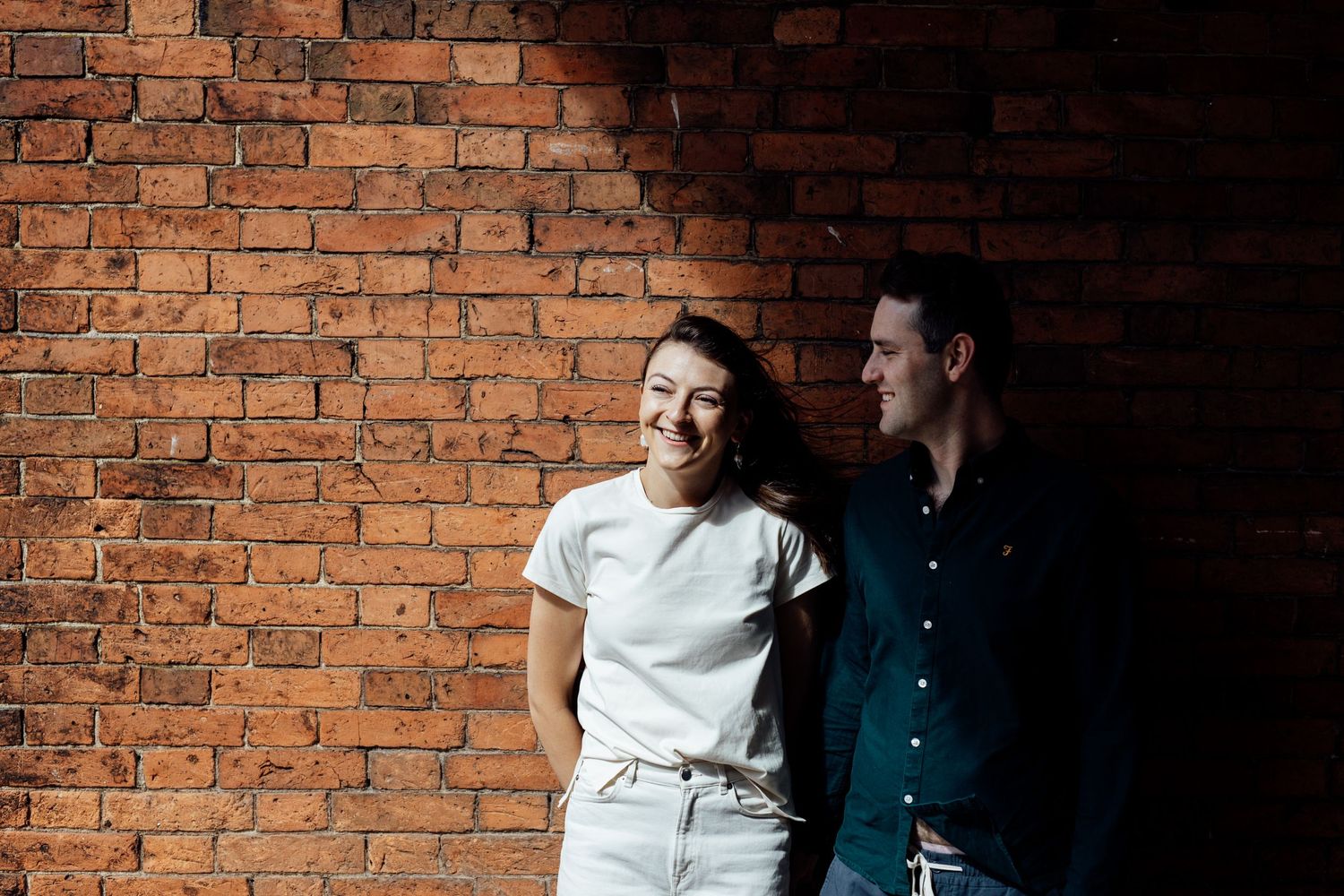A couple poses against a rustic brick wall with dramatic lighting and shadows.