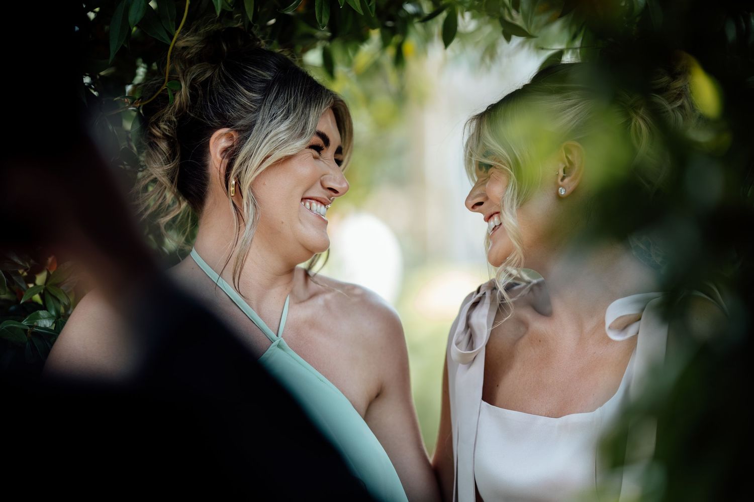 Two brides share a joyful smile among lush green foliage on their wedding day.
