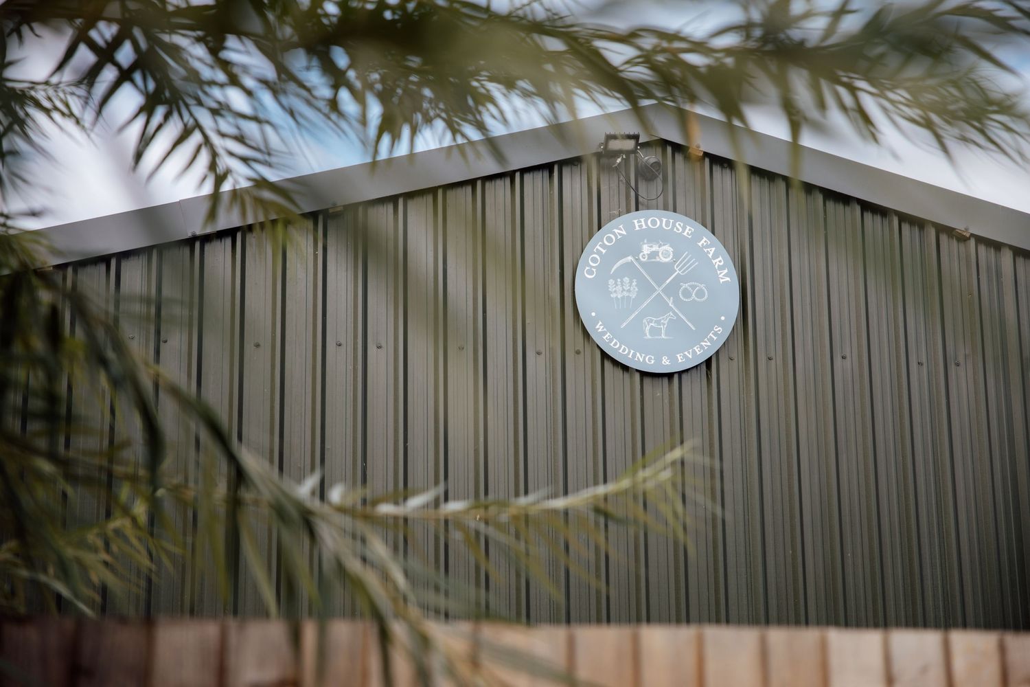 A circular logo sign mounted on a dark green corrugated metal barn wall surrounded by green tree branches.