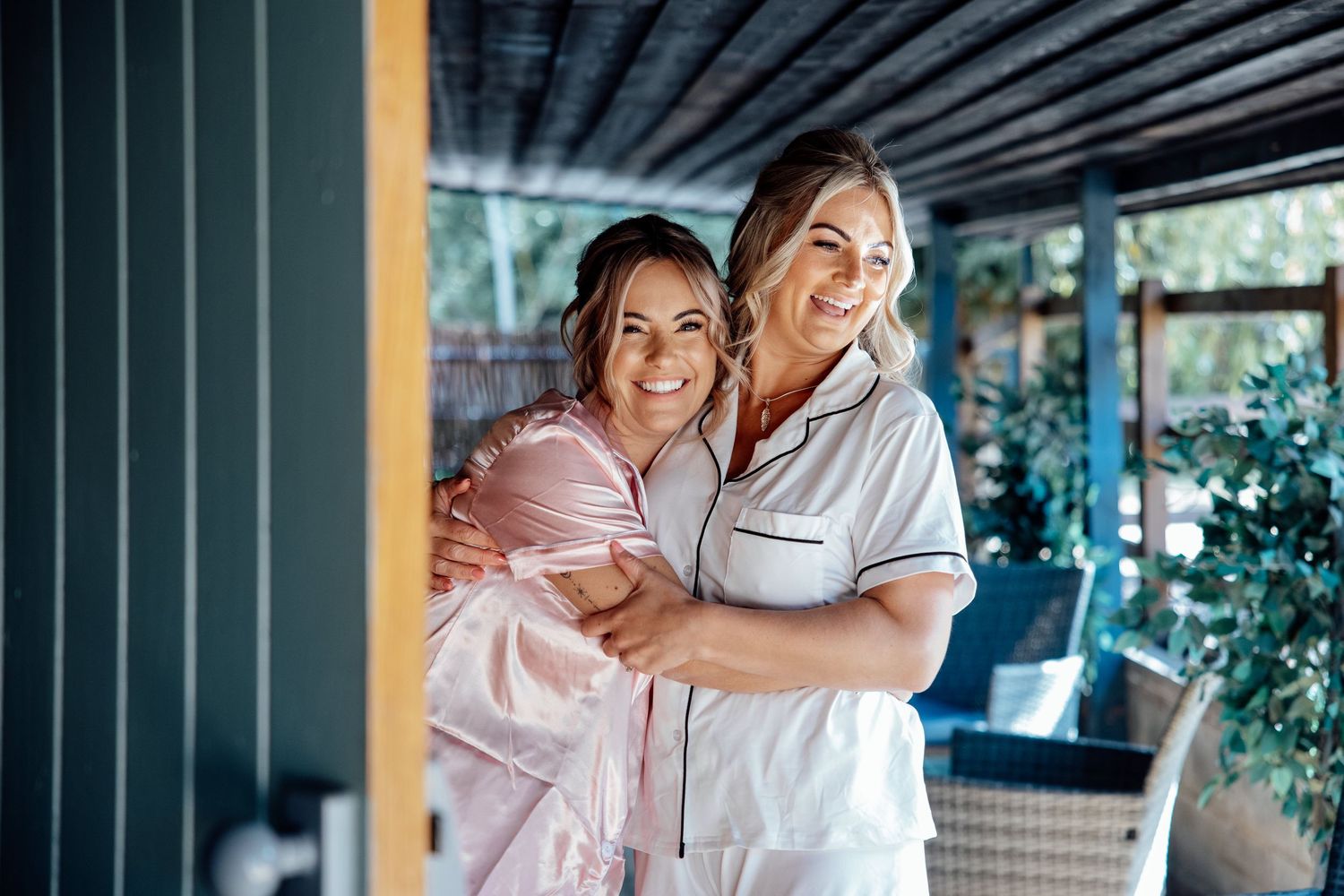 Two women in white and pink pajamas smiling and hugging on a wedding morning.