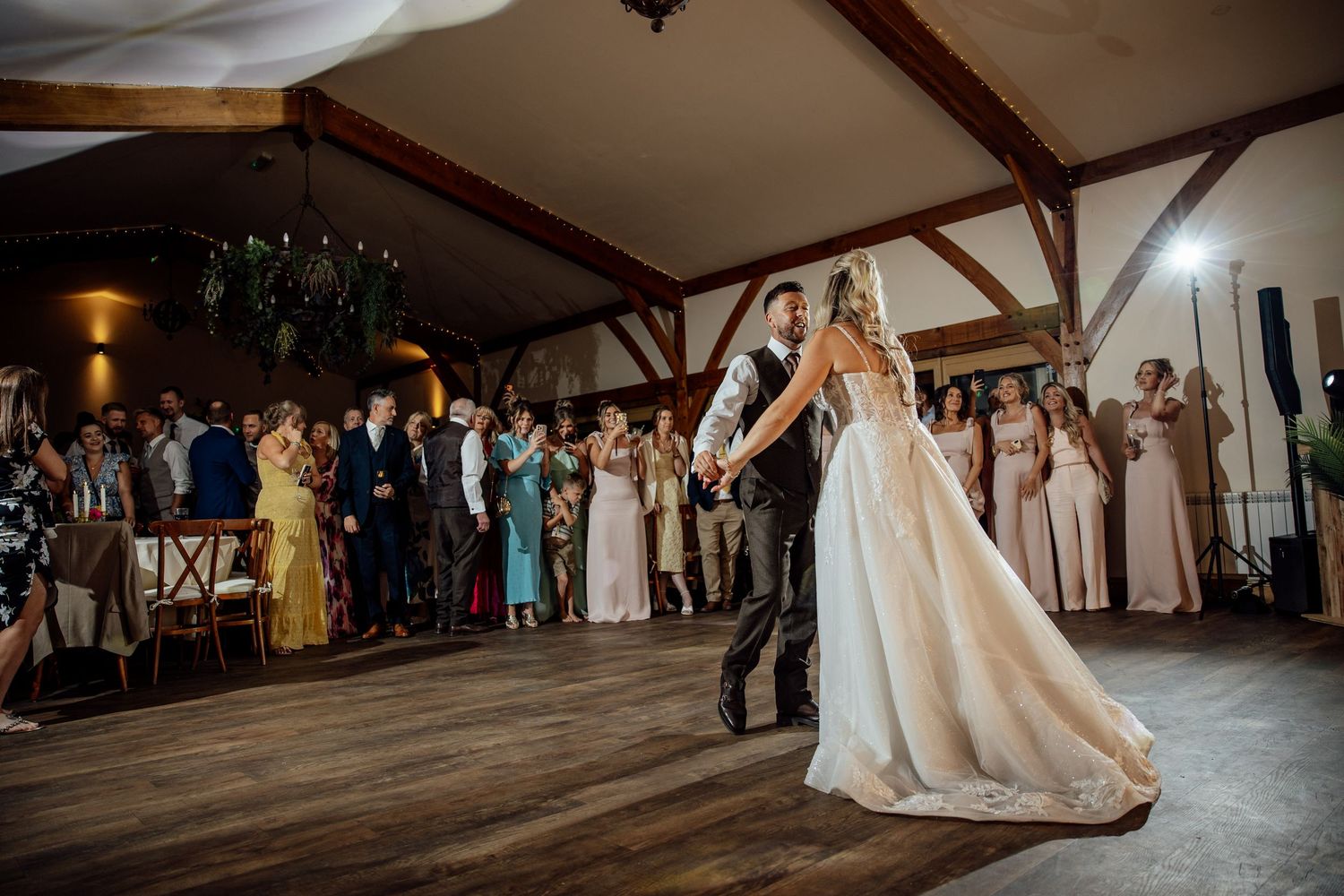 Bride and groom share their first dance in a rustic barn venue while wedding guests applaud around them.