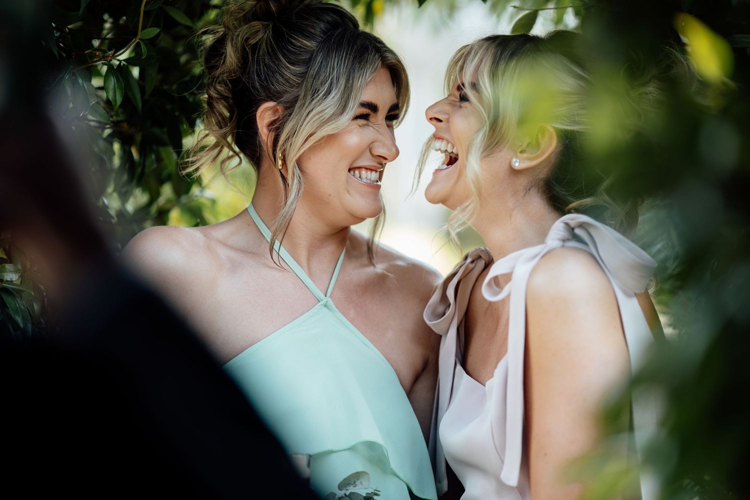Two women in elegant dresses laugh joyfully together outdoors, framed by lush green leaves.