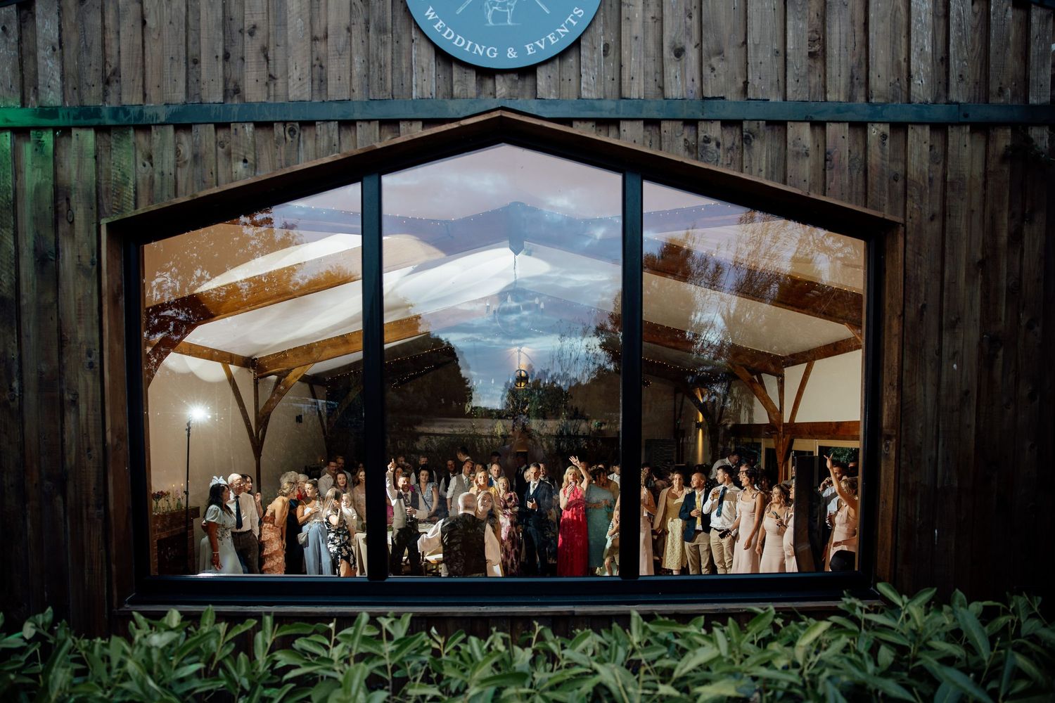 Wedding party guests gathered inside a rustic barn venue, captured through a large triangular window at night.