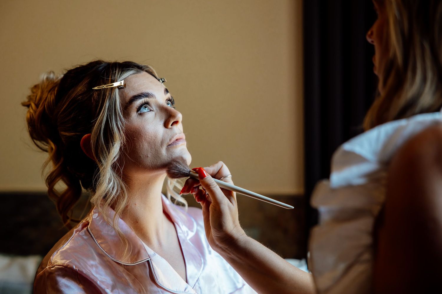 A makeup artist applies foundation to a blonde bride's face with a brush during wedding preparations.