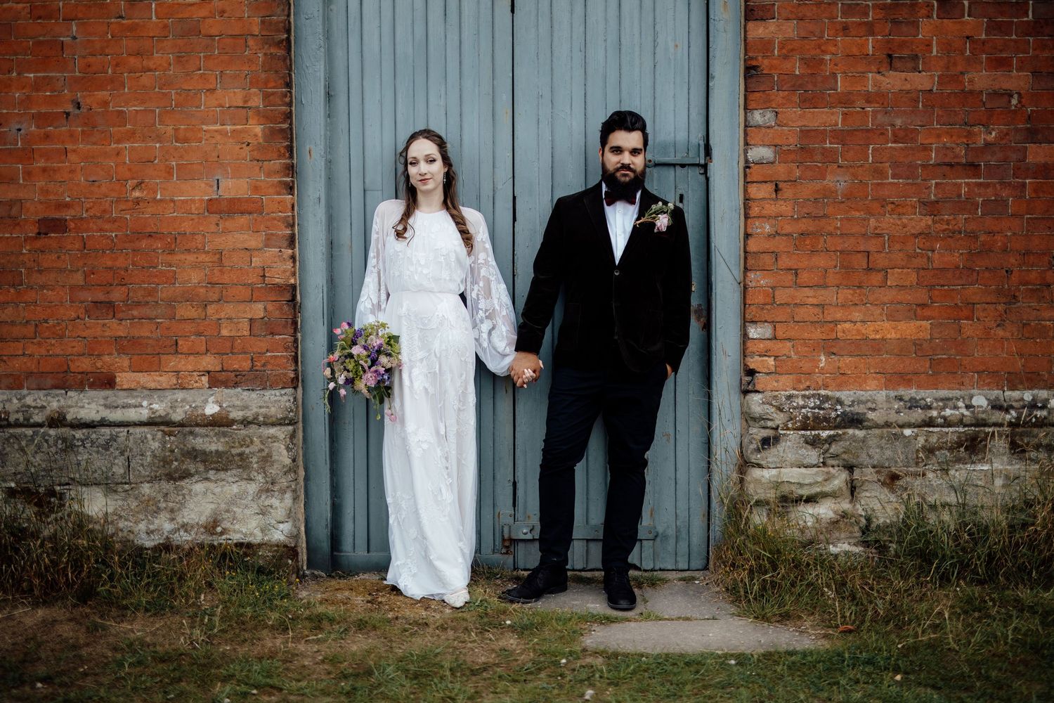 Wedding couple poses against weathered brick wall and vintage blue door in romantic setting.