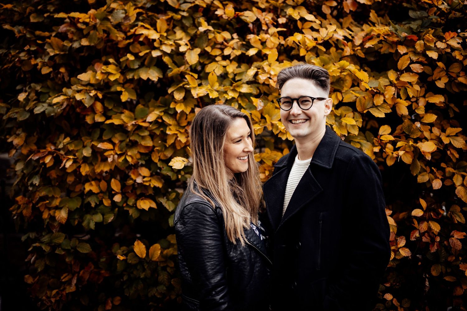 A couple stands against vibrant fall foliage in golden autumn lighting.