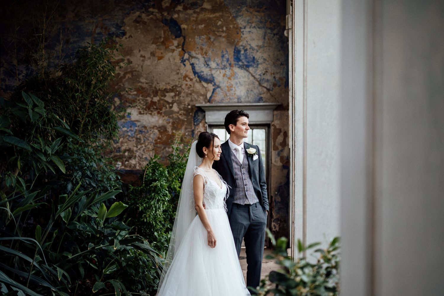 Wedding couple poses against weathered wall with indoor plants.