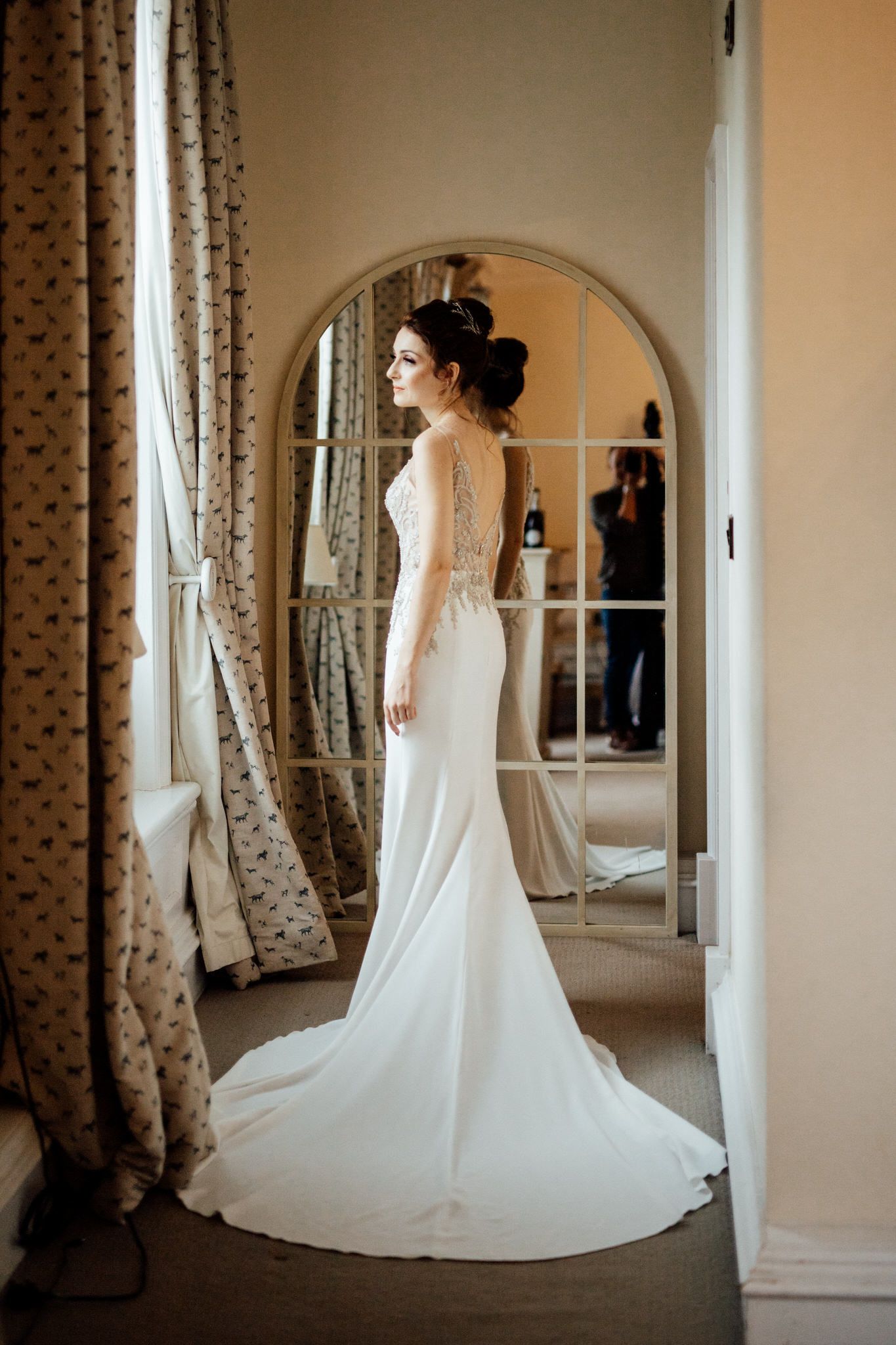 Elegant white wedding dress displayed in room with patterned curtains and arched doorway.