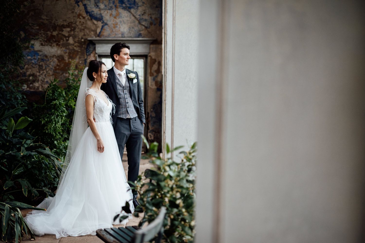 Wedding couple poses against white wall in rustic indoor setting with natural light streaming in.