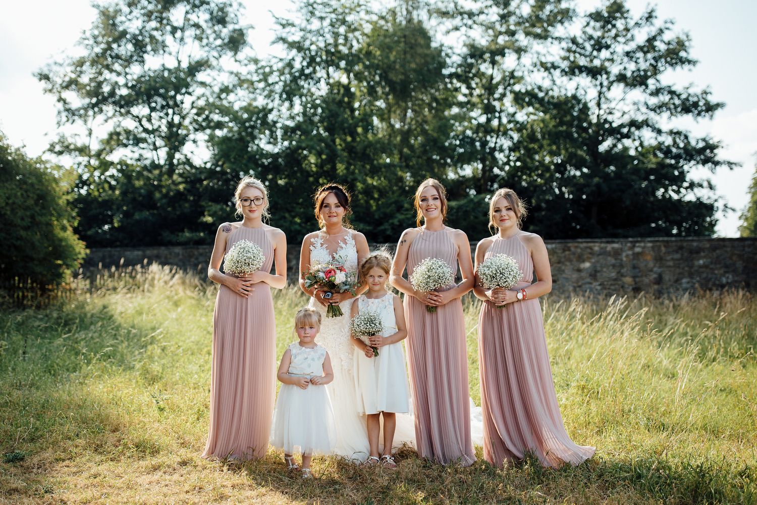Bridesmaids in blush pink dresses standing together in a sunny field.