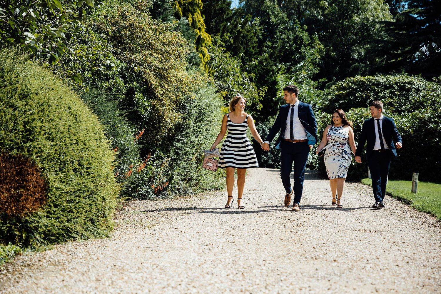 Wedding party walking along a scenic tree-lined gravel path outdoors.