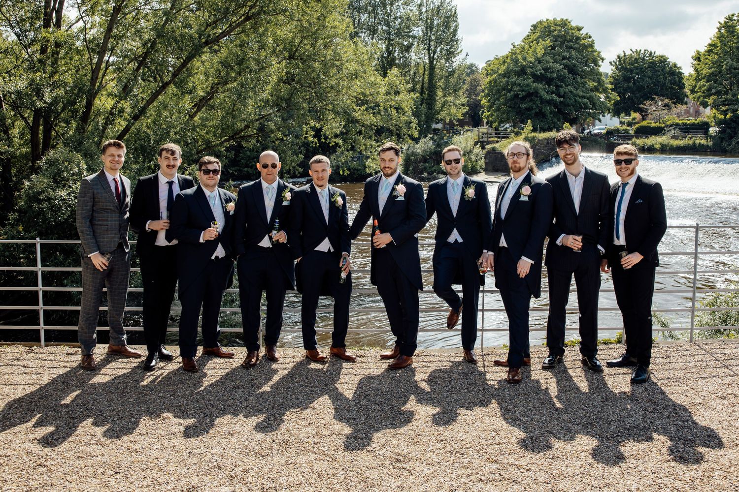 Groomsmen lined up casting shadows on sunny waterfront path.