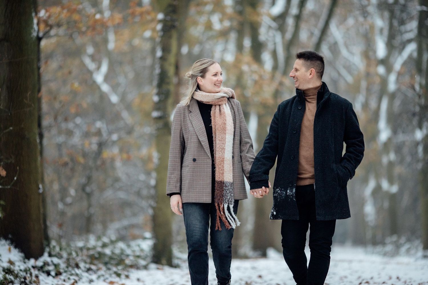 Couple walking through snow-covered winter woods wearing coats.