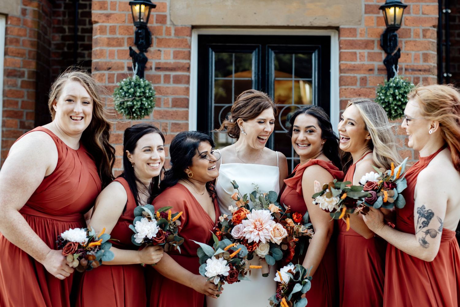 Wedding party in red dresses poses outside brick venue with floral arrangements.