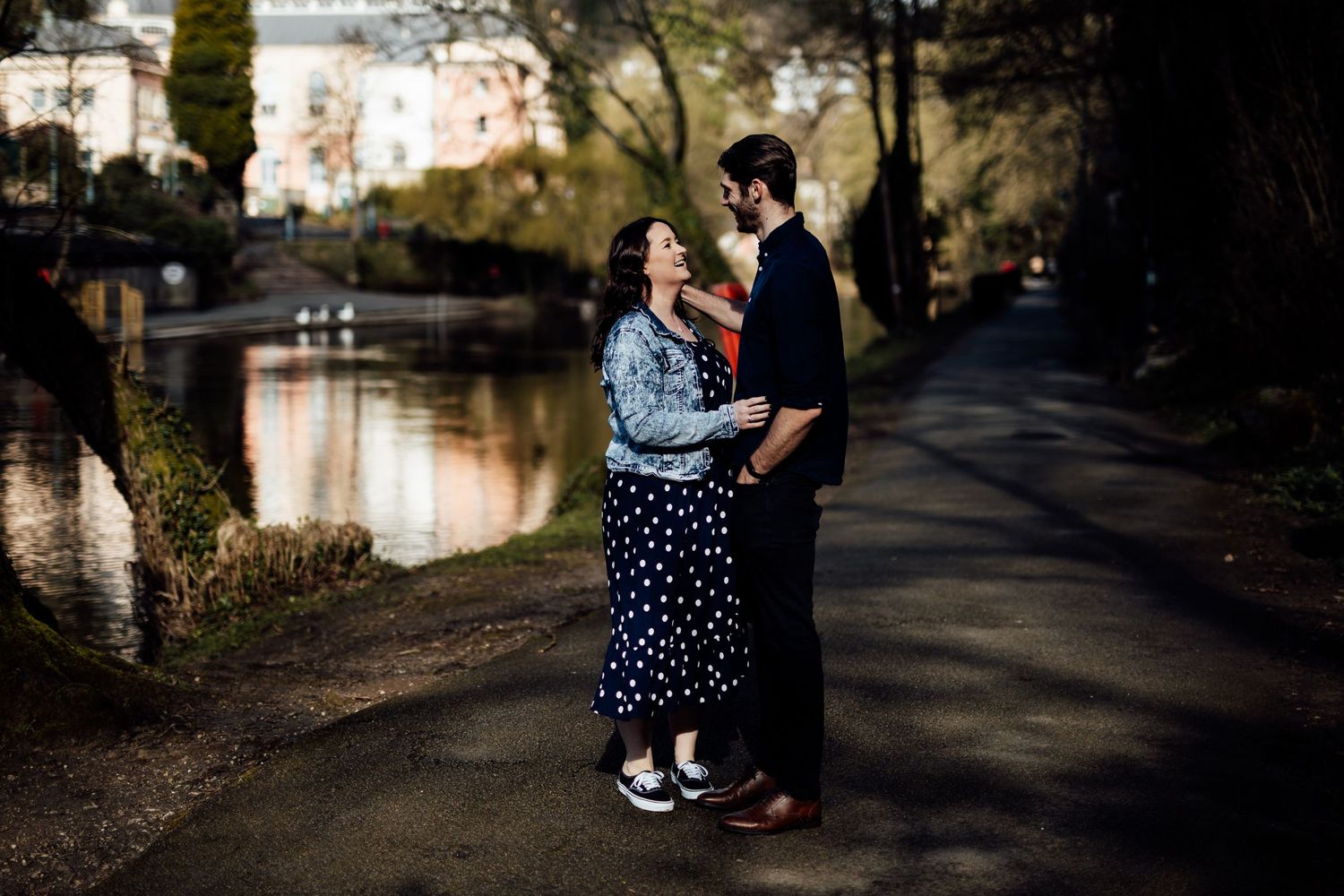 A romantic couple embraces by a canal at sunset wearing casual attire with polka dot dress and dark clothing.