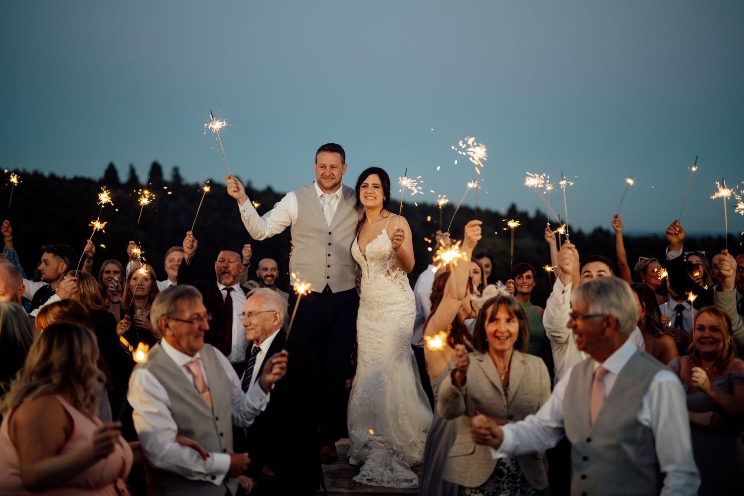 Wedding guests gather outdoors at dusk holding sparklers creating a magical atmosphere.
