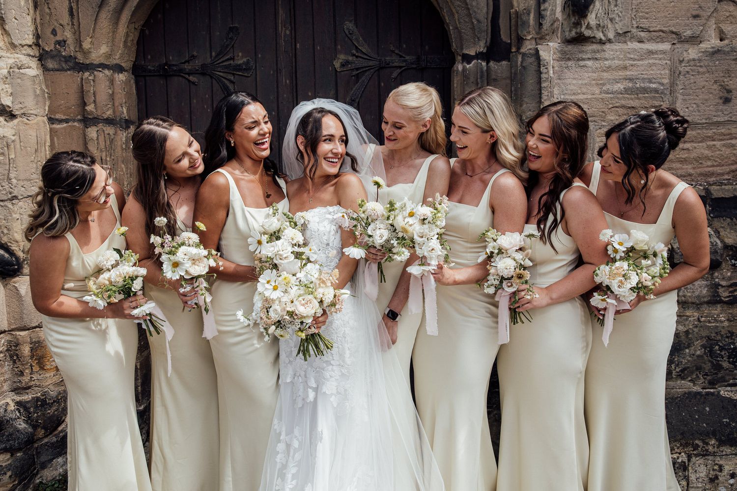Bridesmaids in cream-colored dresses hold white bouquets against a stone wall.