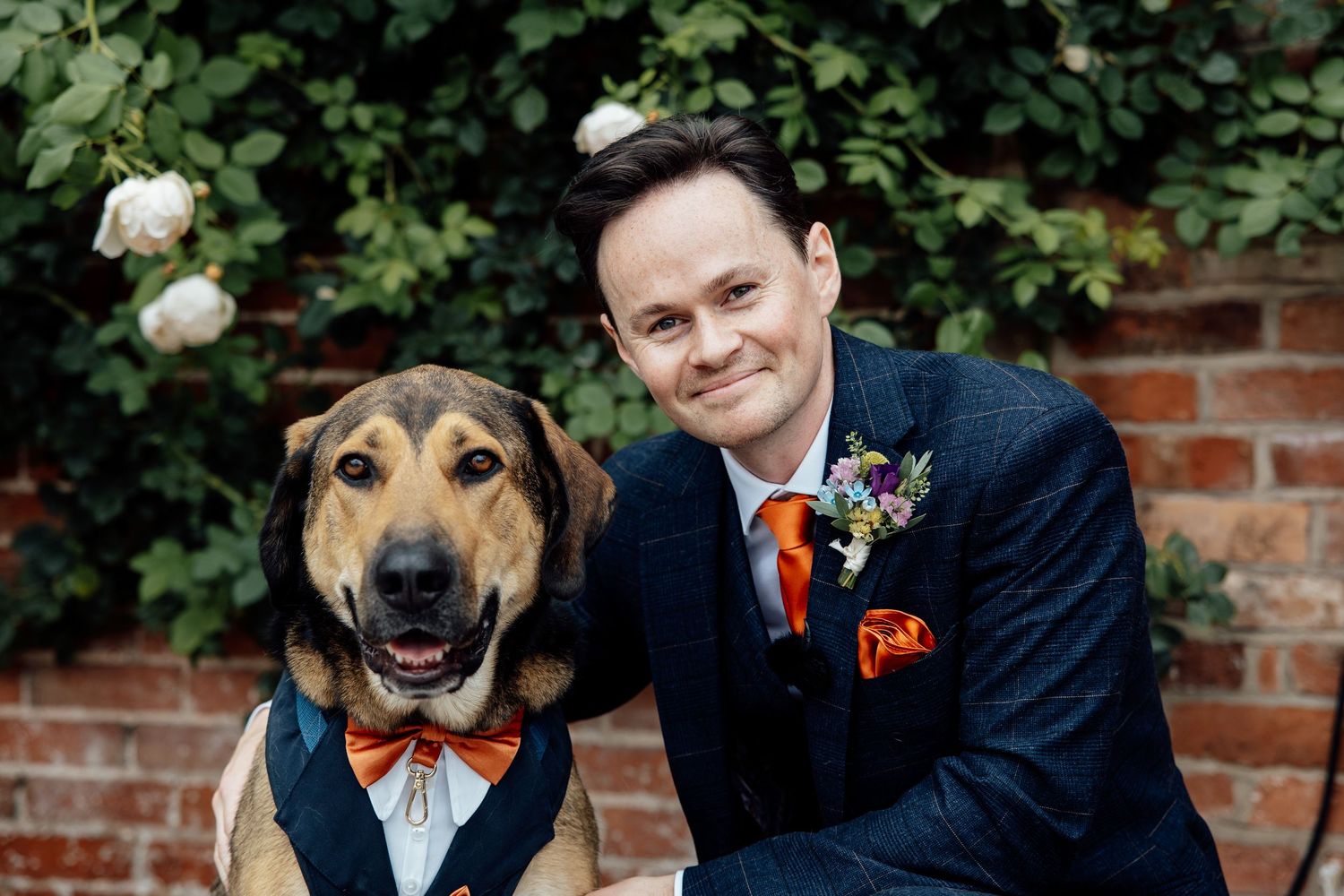 Happy dog wearing bow tie poses with wedding party in garden setting.