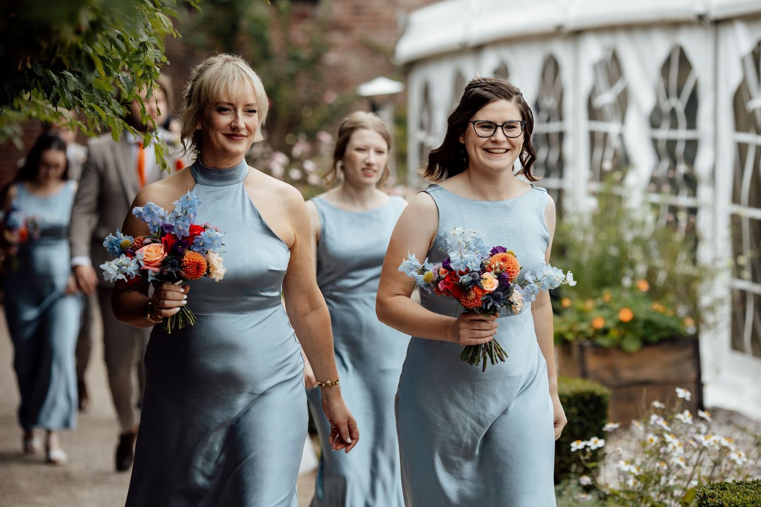 Bridesmaids in blue dresses carry vibrant bouquets while walking through a garden setting.