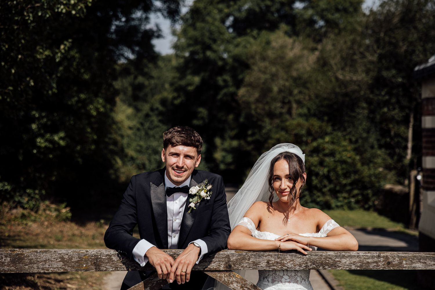 A bride and groom sit casually on a stone wall surrounded by trees on their wedding day.
