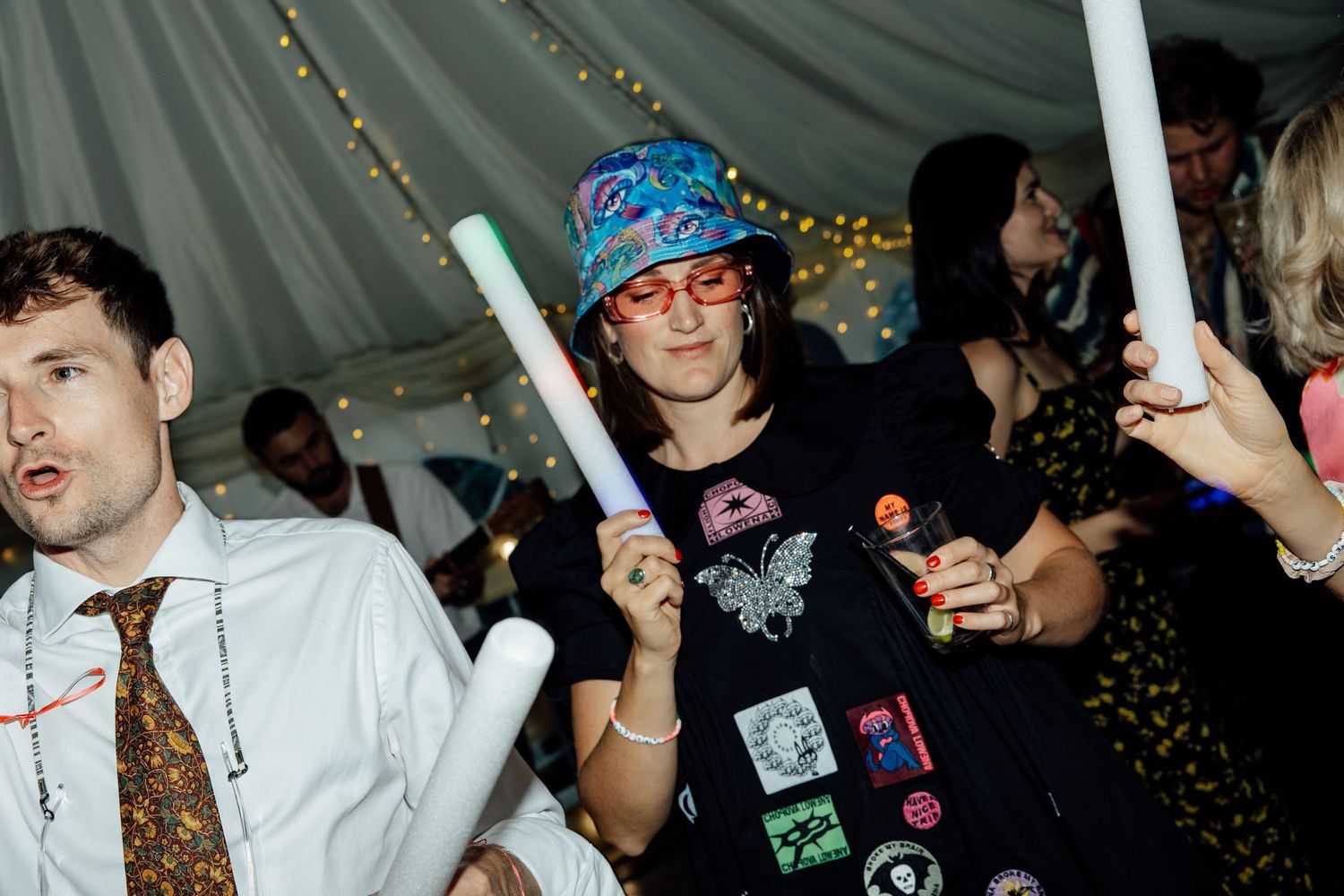Wedding reception party guests dancing and holding glow sticks under a tent.