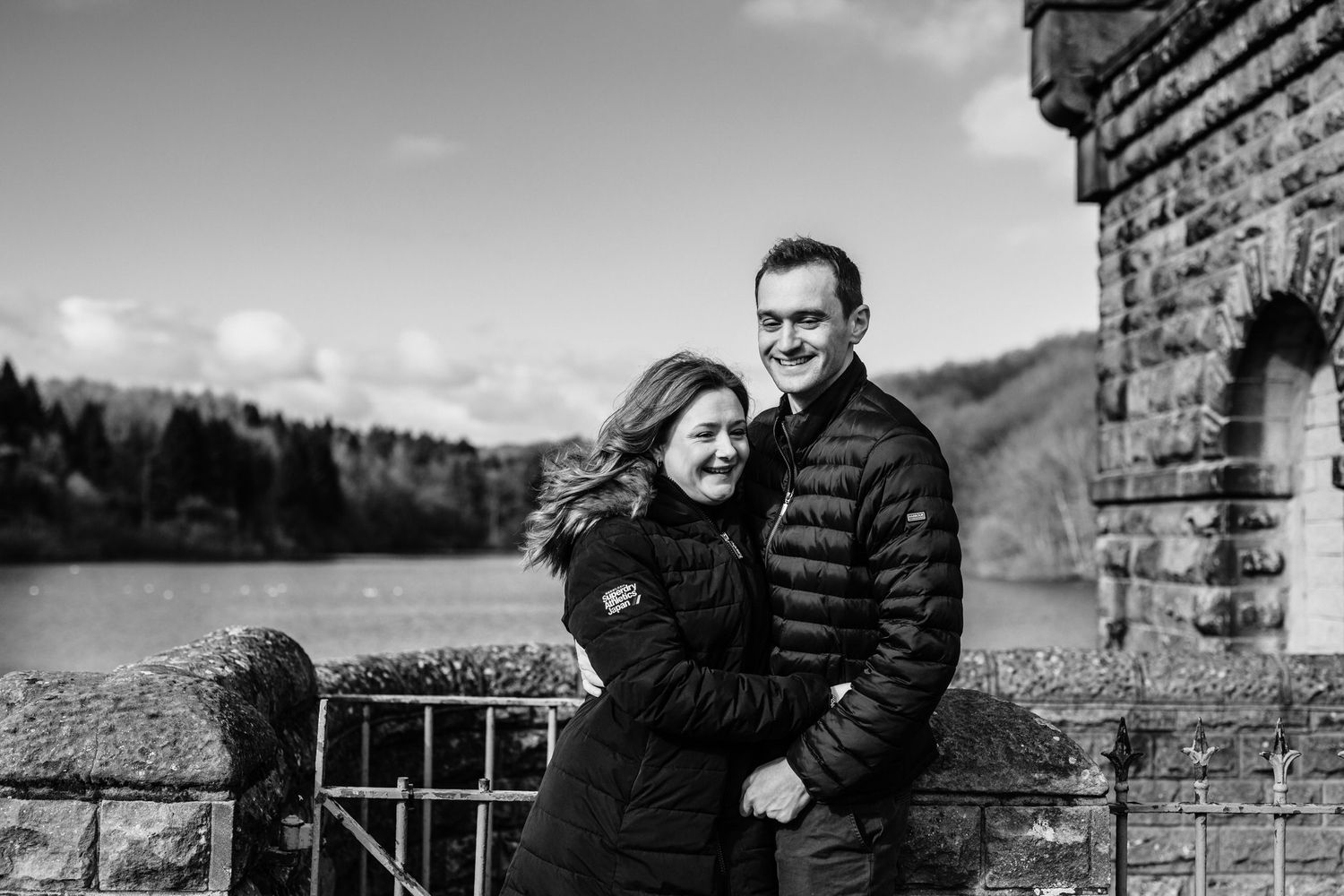 Couple laughing together against rustic stone wall backdrop on cloudy day.