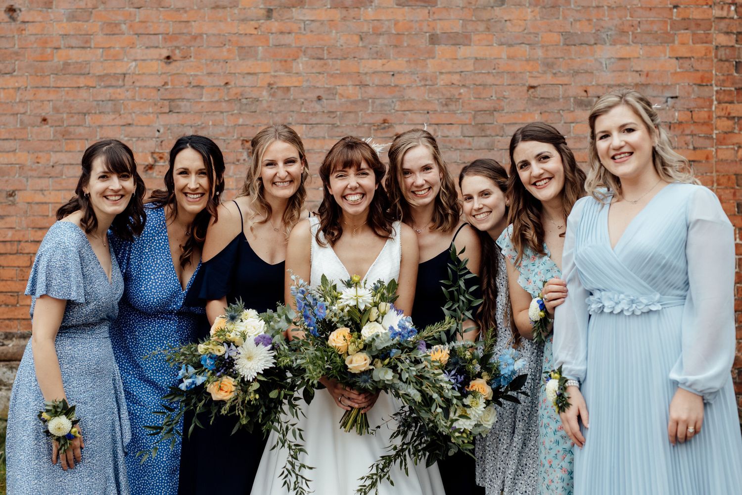 Bridesmaids wearing light blue dresses pose together against a rustic brick wall with floral bouquets.