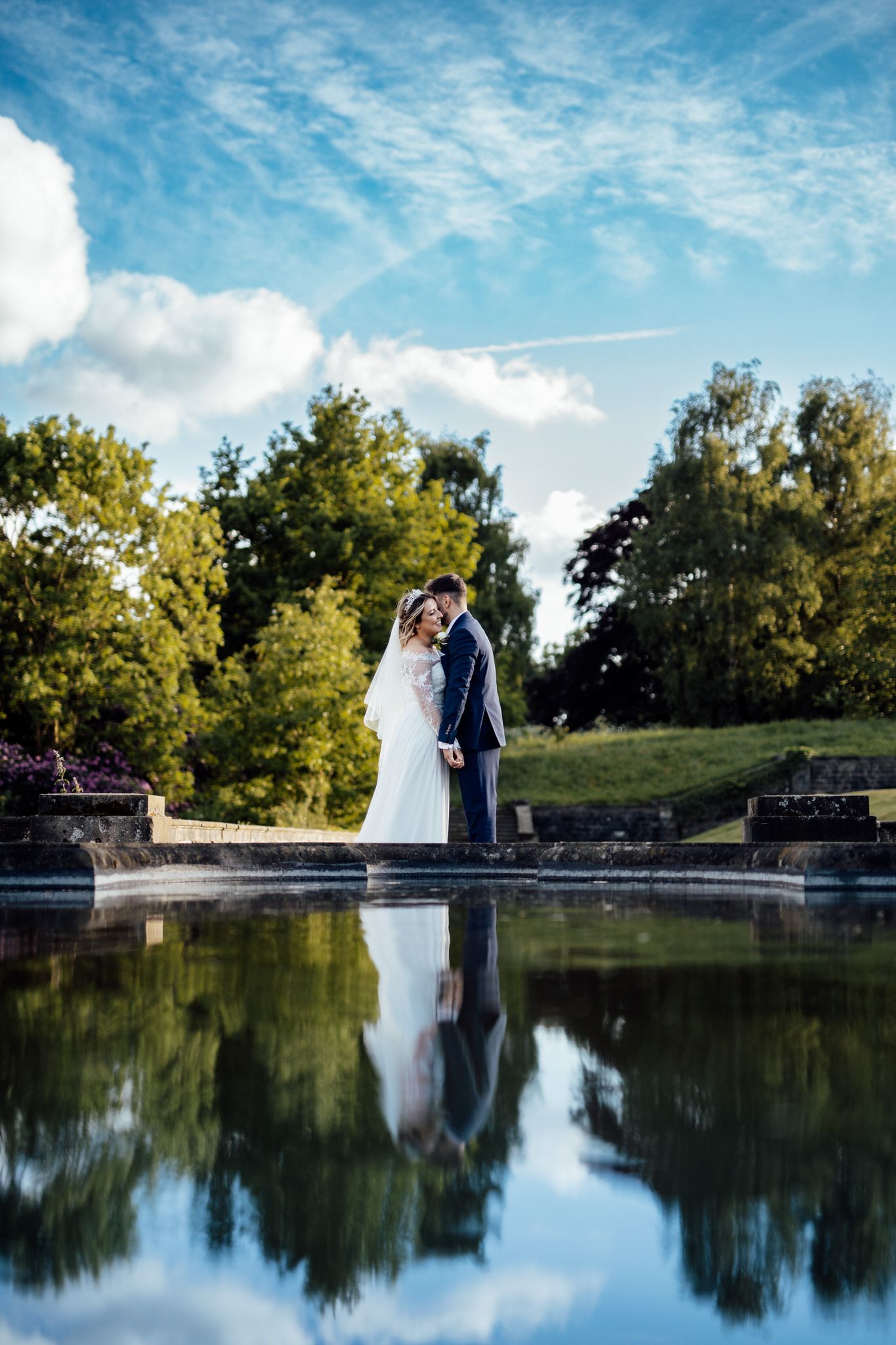 Wedding couple reflected in still water with blue sky and trees in background.
