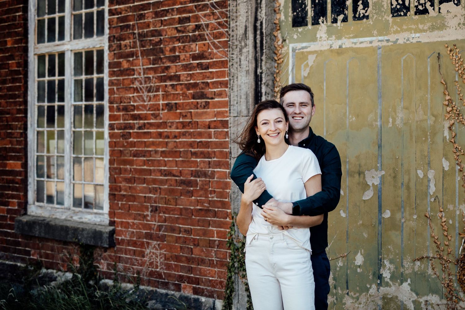 A couple poses playfully against weathered brick walls and vintage windows.