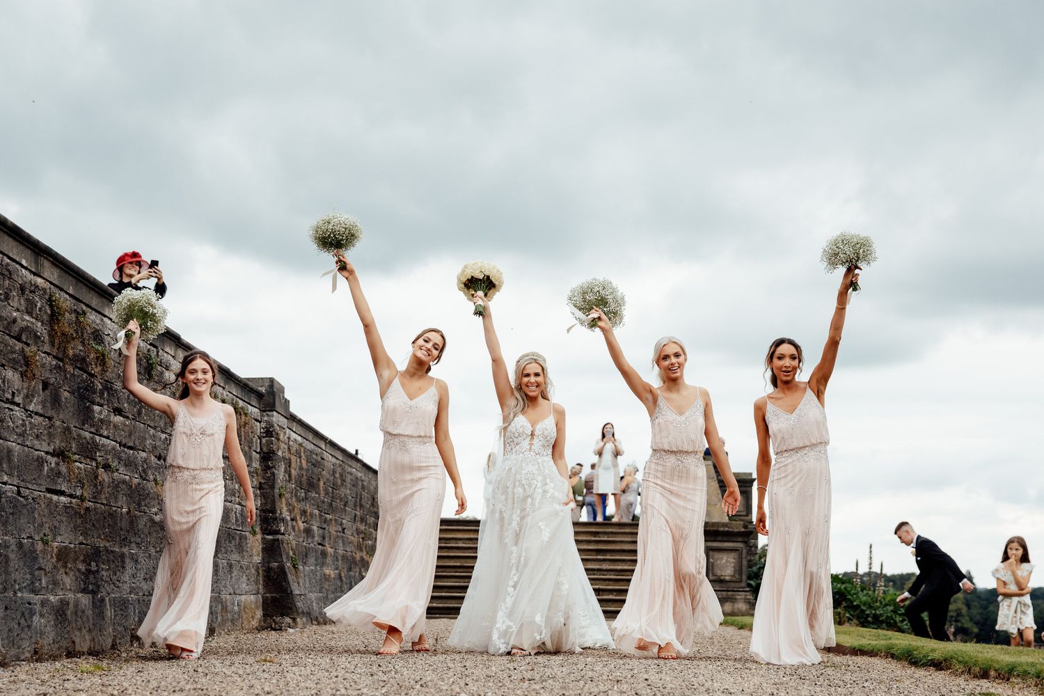 Bridesmaids in white dresses jump joyfully together on a stone wall during a wedding photoshoot.