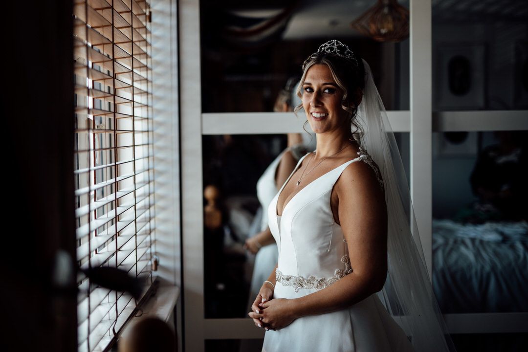 Bride in white dress standing by window with dramatic lighting.