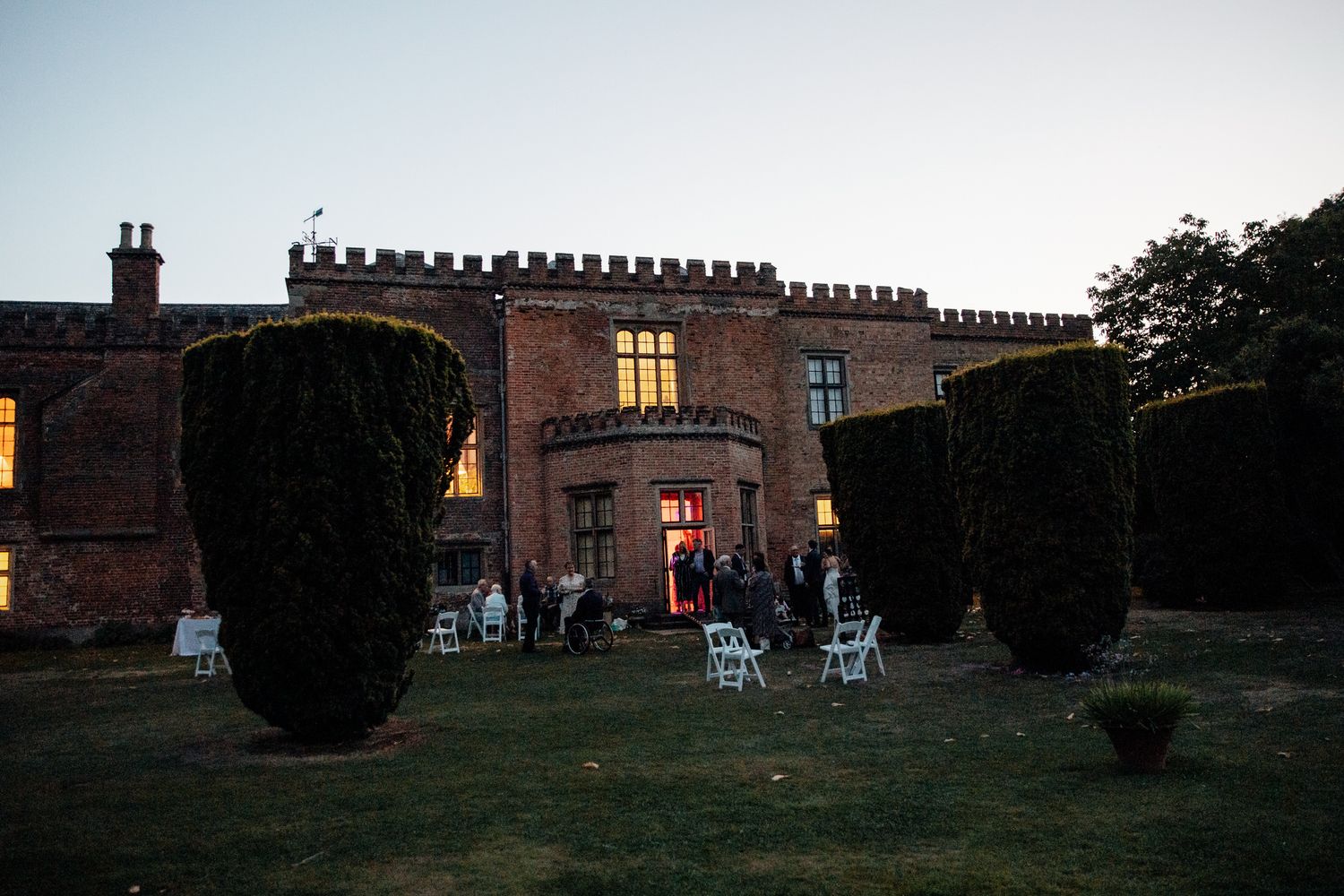 Historic brick mansion at dusk with illuminated windows and garden furniture on the lawn.