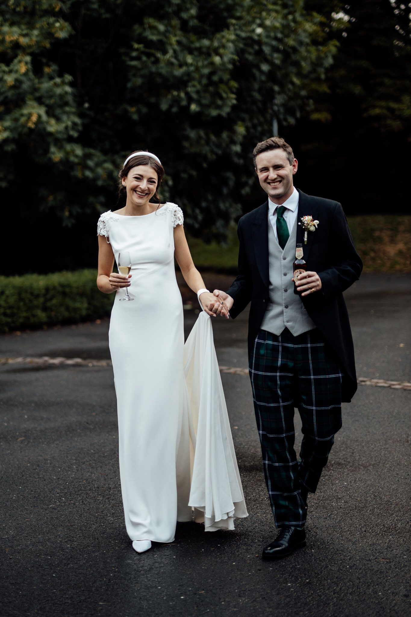 Bride in elegant white dress walks with groom wearing traditional Scottish kilt.