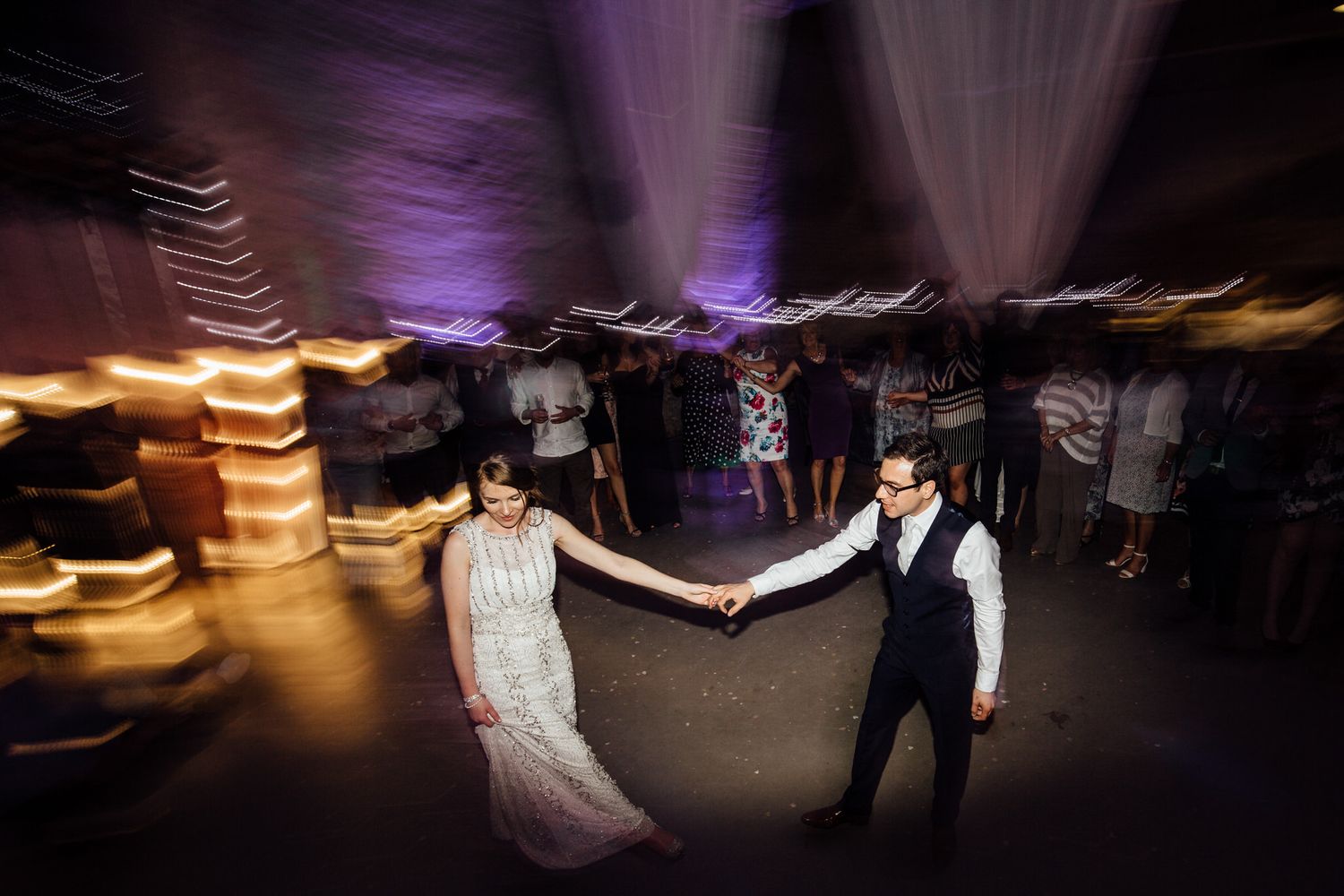Motion-blurred dancers at wedding reception with white drapes and ambient lighting.